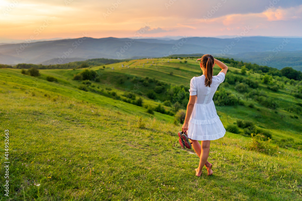 Holiday in the country with a Caucasian woman dressed in a white dress sitting barefoot in the grass and enjoying the beautiful landscape of nature with green hills and dense forest at sunset. Holiday in the country with a Caucasian woman dressed in a white dress sitting barefoot in the grass and enjoying the beautiful landscape of nature with green hills and dense forest at sunset.