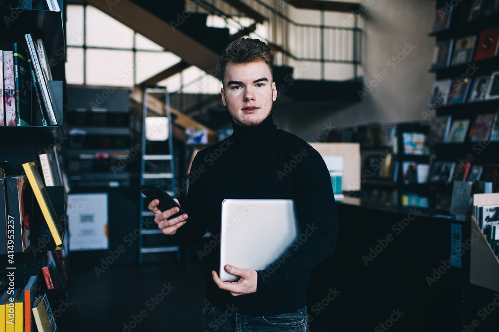 Pensive male student with laptop and smartphone standing in library