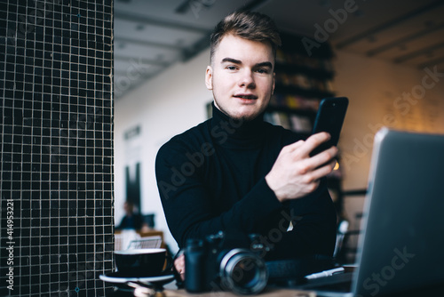 Photographer browsing internet on smartphone near laptop in cafeteria