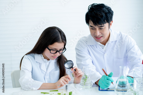 Young Caucasian scientist child girl look through a piece of flower and see more details with magnifying glass with guide and support from Asian teacher in laboratory or classroom.