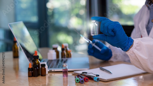 Doctors hold a vial of COVID-19 vaccine in a blue rubber glove in a close-up laboratory. Study the concept of coronavirus vaccination. On the table there were bottles of pills and a laptop with a note © ArLawKa