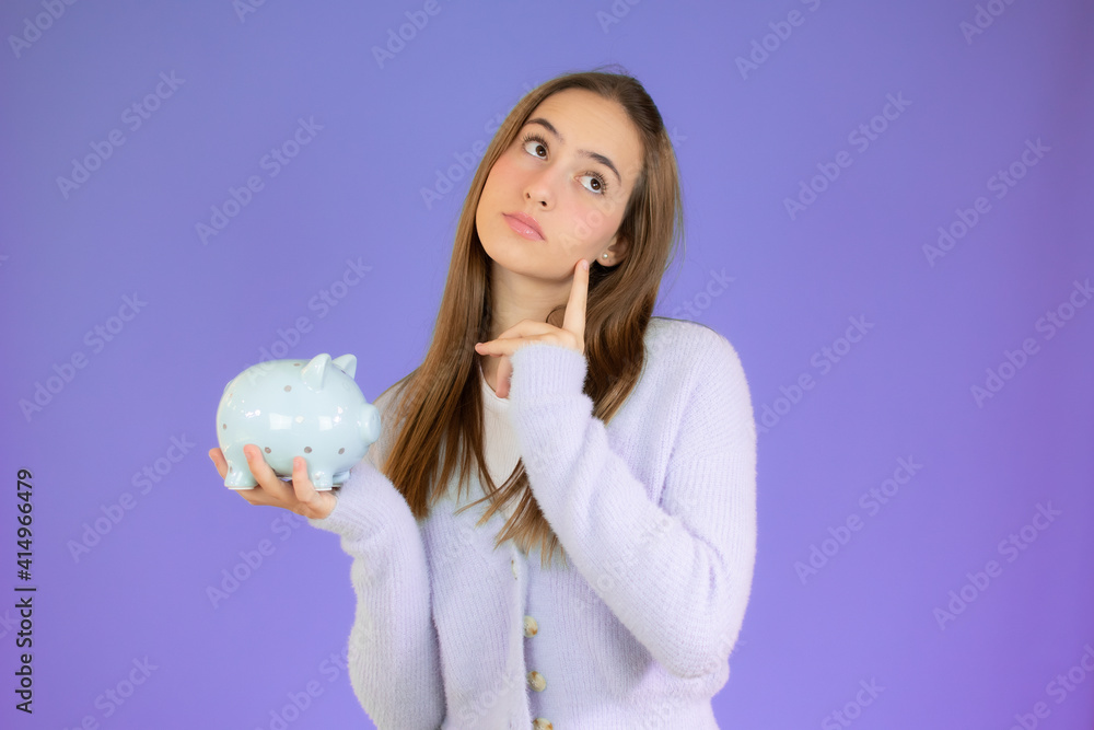 Portrait of a pensive casual girl holding a piggy bank looking away at copy space isolated over purple background Portrait of a pensive casual girl holding a piggy bank looking away at copy space isolated over purple background
