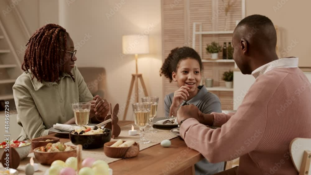 Steadicam shot of happy African family sitting at festive dinner table, cheerful daughter smiling, talking, telling her interesting story to father and mother looking at parents Steadicam shot of happy African family sitting at festive dinner table, cheerful daughter smiling, talking, telling her interesting story to father and mother looking at parents