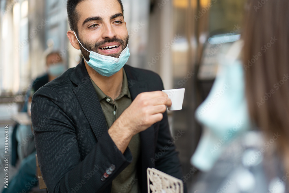 Handsome guy in suit with lowered face mask talking and drinking espresso coffee in an outdoor cafe Handsome guy in suit with lowered face mask talking and drinking espresso coffee in an outdoor cafe