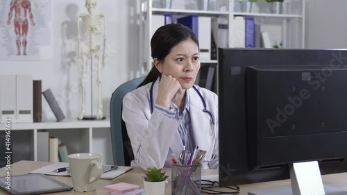 asian physician working on the computer is propping her face, staring at screen and tapping her temples with finger while thinking hard on her office.