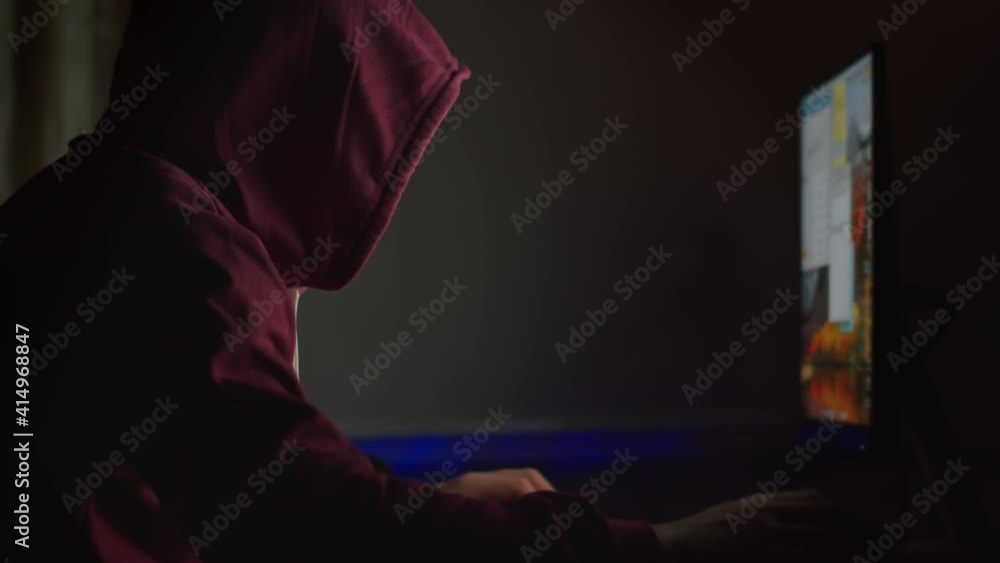 Unidentified man in a hood sits at a computer monitor at night, cracking a password, cyber attack Unidentified man in a hood sits at a computer monitor at night, cracking a password, cyber attack