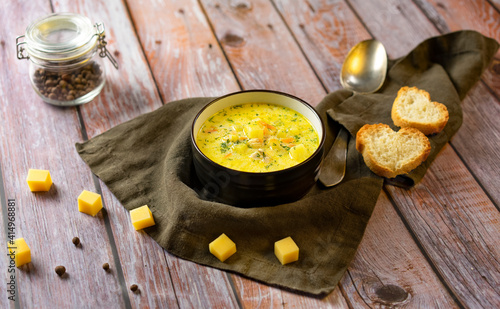 Cheese yellow soup in a dark plate. Pieces of cheese and bread around the plate. On a textured wooden background. Horizontal orientation.