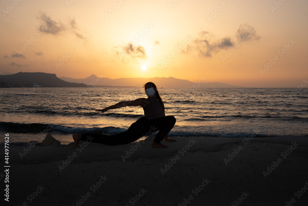 Fototapeta premium Silhouette of unrecognized young woman with protective surgical face mask performs yoga stretching exercises at the beach at dusk during coronavirus pandemic.