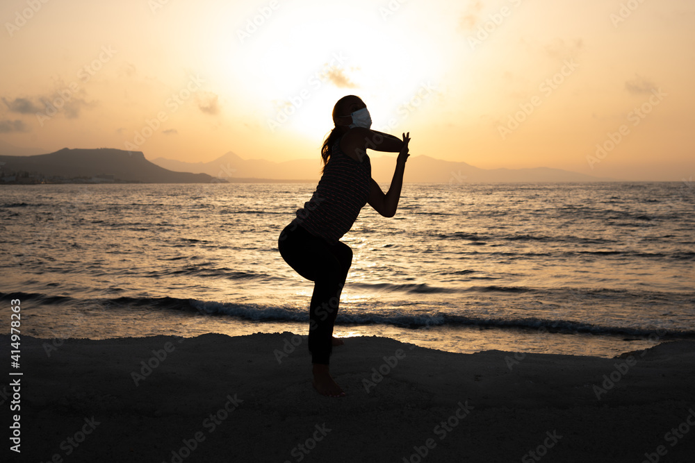 Silhouette of unrecognized young woman with protective surgical face mask performs yoga stretching exercises at the beach at dusk during coronavirus pandemic.