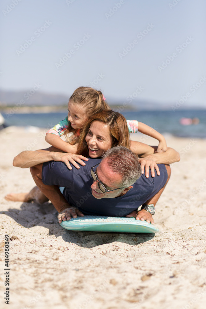 Allegra famiglia composta da padre, madre e figlia si diverte giocando con con una tavola da surf in spiaggia al mare Allegra famiglia composta da padre, madre e figlia si diverte giocando con con una tavola da surf in spiaggia al mare