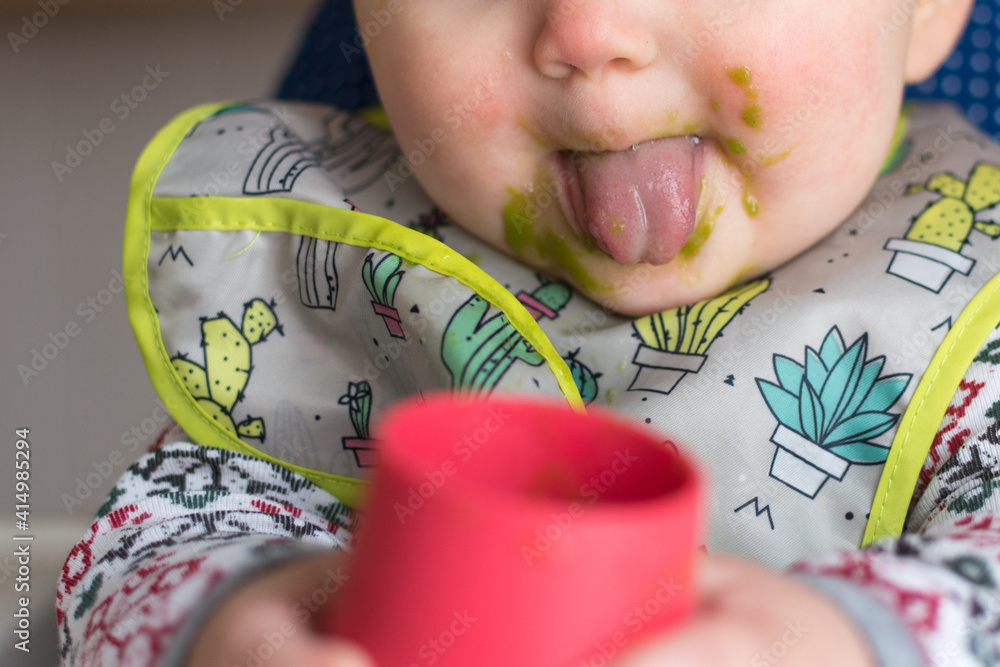 Baby with a tongue tie drinking green vegetable smoothie; tongue sticking out with messy face