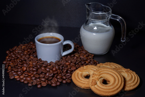 Fototapeta Naklejka Na Ścianę i Meble -  White coffee cup with a coffee drink inside and surrounded by roasted coffee beans of the Robusta variety, a glass jug with milk inside and a donut cake. Breakfast concept