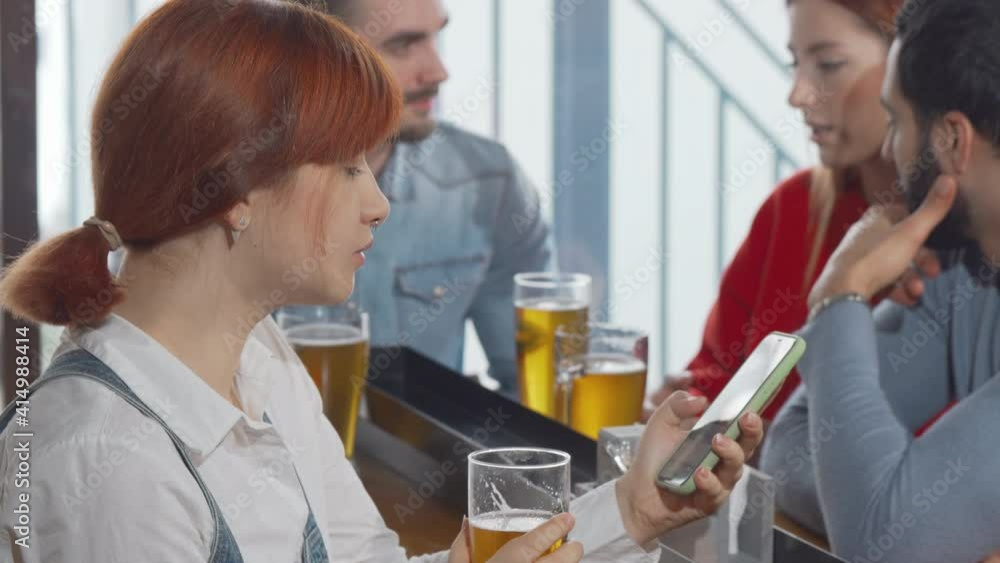 Young woman using her smart phone while drinking beer at the pub. Relaxed woman sipping delicious beer, browsing online on her phone. Technology, social media concept Young woman using her smart phone while drinking beer at the pub. Relaxed woman sipping delicious beer, browsing online on her phone. Technology, social media concept