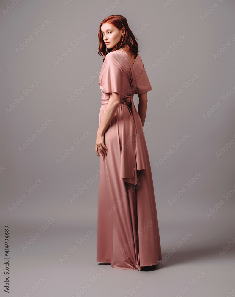 Bridesmaid's look in dust pink. Young ginger woman in summer transformer evening dress and high heel shoes on grey background at studio. Gorgeous sexy lady. Bridesmaid's look in dust pink. Young ginger woman in summer transformer evening dress and high heel shoes on grey background at studio. Gorgeous sexy lady.
