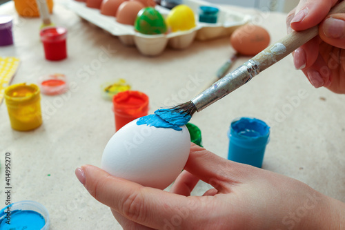 Process of handmade decoration of Easter egg in blue colour paint at home and folowing traditions. Woman's hands close-up holding brush and multicolored jars of paint in background. photo