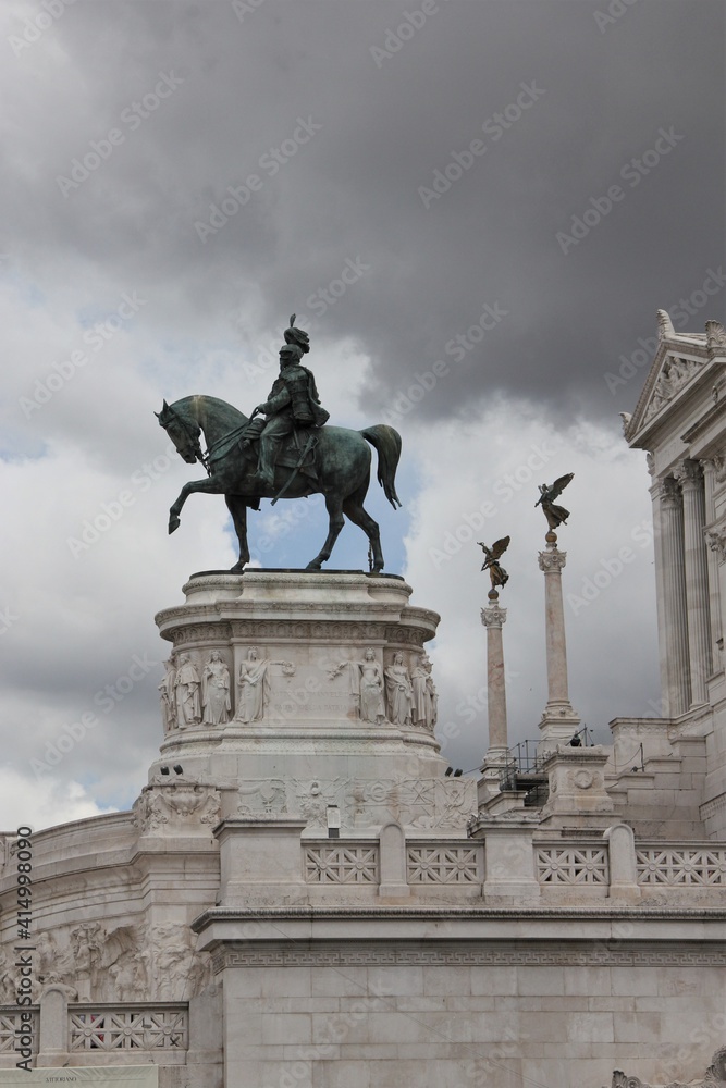 Equestrian green patina bronze statue of Victor Emmanuel II in front of the white marble clad Altare della Patria building against a dramatic dark storm cloud sky, Rome 
