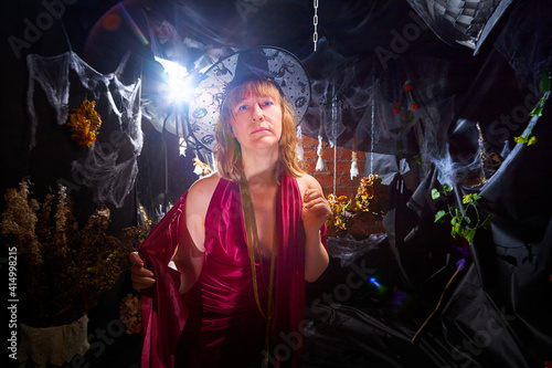 Blonde witch in red dress and black hat in Halloween decoration indoors.