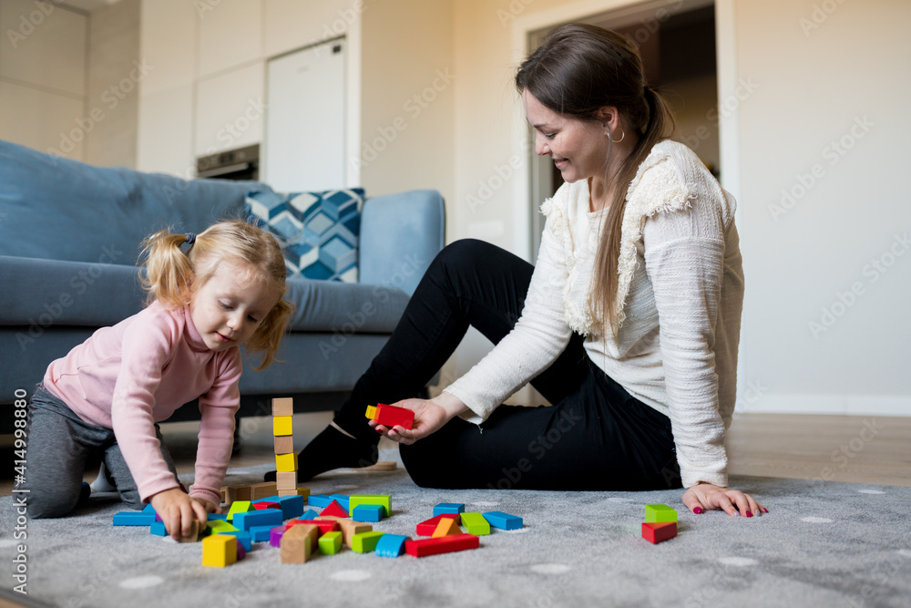 Naklejka premium Mother and daughter playing wooden cubes on the floor of the living room at home. Activities with children inside, early childhood development
