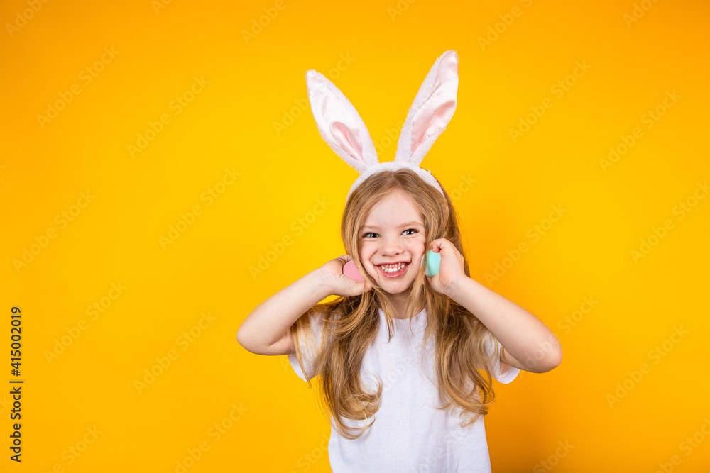 Baby girl with bunny ears holding Easter eggs in her hands near her face on a yellow background