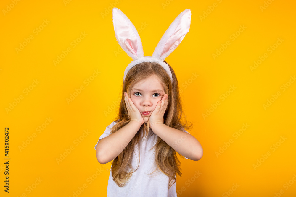 Portrait of a surprised Easter girl with bunny ears on a yellow background holding her face with her hands