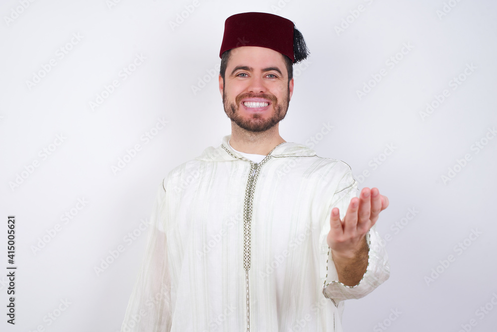 young handsome Caucasian man wearing Arab djellaba and Fez hat over white wall smiling cheerful offering palm hand giving assistance and acceptance. young handsome Caucasian man wearing Arab djellaba and Fez hat over white wall smiling cheerful offering palm hand giving assistance and acceptance.