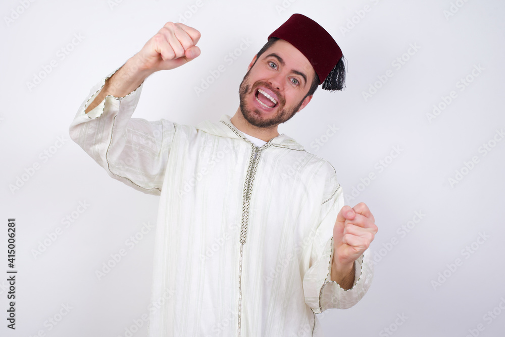 Naklejka premium young handsome Caucasian man wearing Arab djellaba and Fez hat over white wall imagine steering wheel helm rudder passing driving exam good mood fast speed