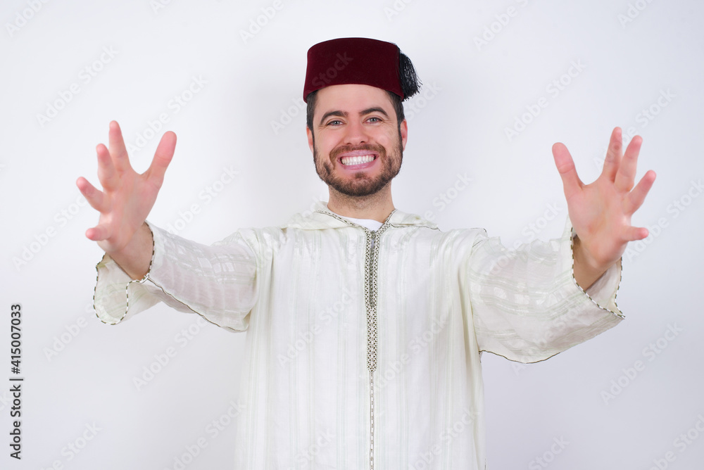 young handsome Caucasian man wearing Arab djellaba and Fez hat over white wall looking at the camera smiling with open arms for hug. Cheerful expression embracing happiness. young handsome Caucasian man wearing Arab djellaba and Fez hat over white wall looking at the camera smiling with open arms for hug. Cheerful expression embracing happiness.