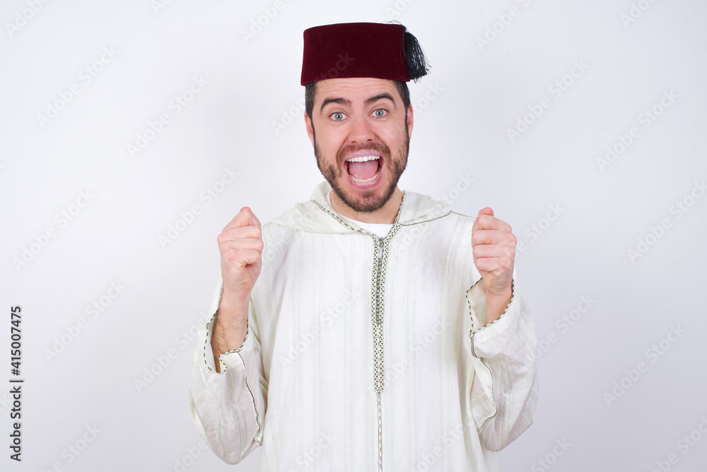 young handsome Caucasian man wearing Arab djellaba and Fez hat over white wall celebrating surprised and amazed for success with arms raised and open eyes. Winner concept. young handsome Caucasian man wearing Arab djellaba and Fez hat over white wall celebrating surprised and amazed for success with arms raised and open eyes. Winner concept.