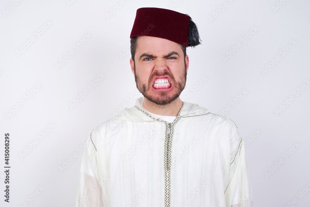 young handsome Caucasian man wearing Arab djellaba and Fez hat over white wall keeps teeth clenched, frowns face in dissatisfaction, irritated because of much duties.