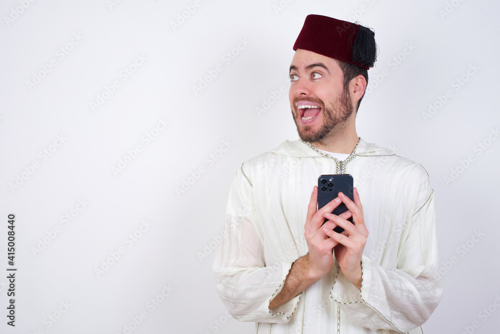 Copyspace photo of young handsome Caucasian man wearing Arab djellaba and Fez hat over white wall stupor with something occurring in social media Copyspace photo of young handsome Caucasian man wearing Arab djellaba and Fez hat over white wall stupor with something occurring in social media