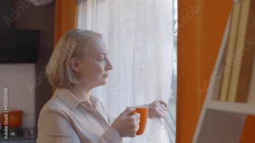 Side view of mature elegant female in glasses enjoying morning coffee standing by the window and smiling.The old woman is looking out the window while drinking coffee at his home.