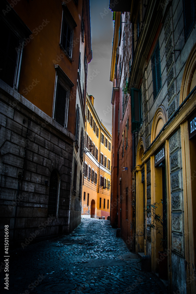Una estrecha y sombría calle de Roma con un edificio iluminado al fondo y con sus tradicionales colores anaranjados en tonos pastel
