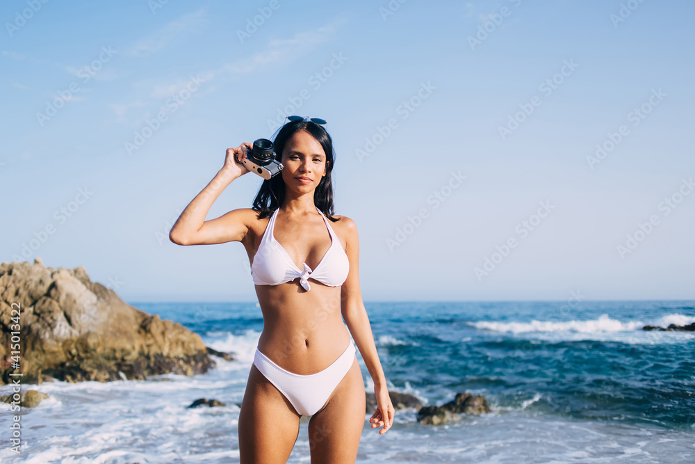 Portrait of charming girl with perfect sexy body and tanned skin looking at camera while testing retro equipment during summer weekend at beach, Latin female photographer recreating near Caribbean sea
