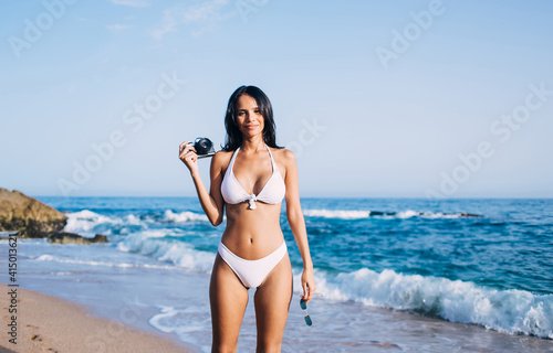 Half length portrait of sexy female photographer dressed in white bikini looking at camera while using retro equipment during sunbathing at beach, beautiful Latin amateur with old fashioned technology