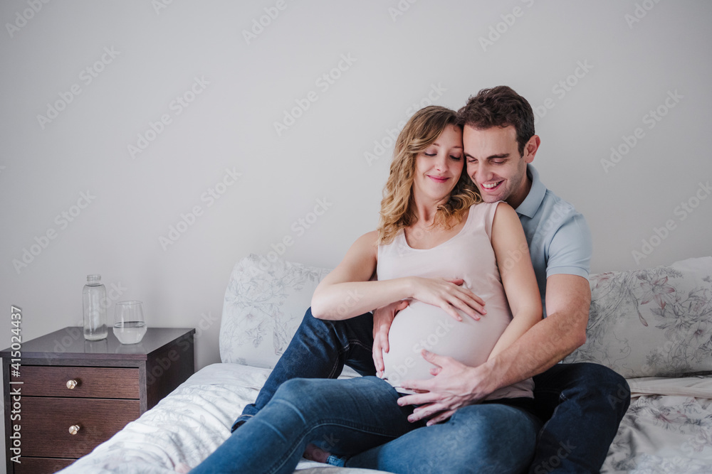 Smiling man touching pregnant wife's stomach while sitting on bed at home Smiling man touching pregnant wife's stomach while sitting on bed at home