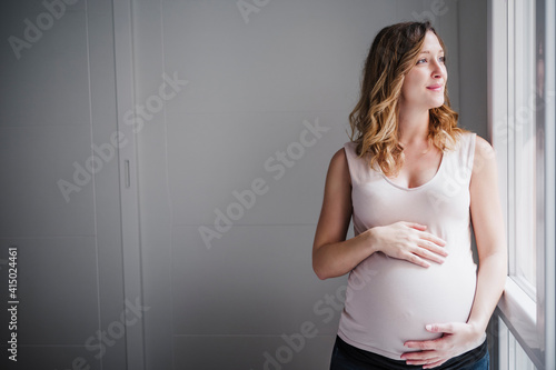 Thoughtful pregnant woman with hands on stomach looking through window at home