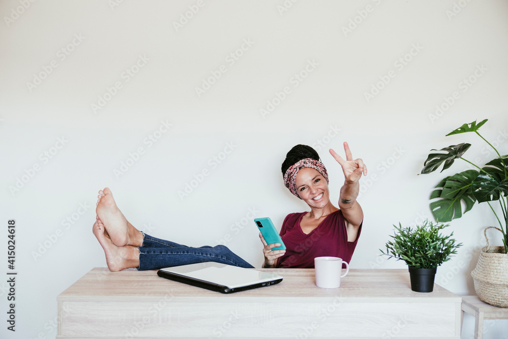 © Eva Blanco/Westend61 - Cheerful female entrepreneur making peace sign while relaxing with feet up at home office