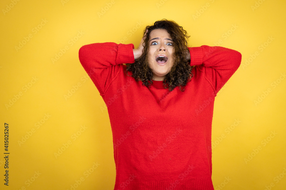 Young african american woman wearing red sweater over yellow background crazy and scared with hands on head, afraid and surprised of shock Young african american woman wearing red sweater over yellow background crazy and scared with hands on head, afraid and surprised of shock