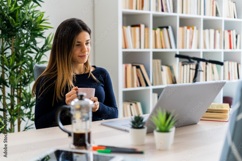 Female freelancer looking at laptop while holding coffee cup in home Female freelancer looking at laptop while holding coffee cup in home