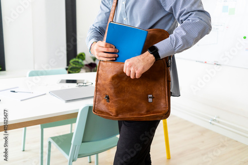 Young businessman putting notepad into shoulder bag in creative office