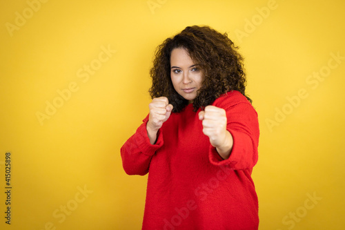 Young african american woman wearing red sweater over yellow background Punching fist to fight, aggressive and angry attack, threat and violence