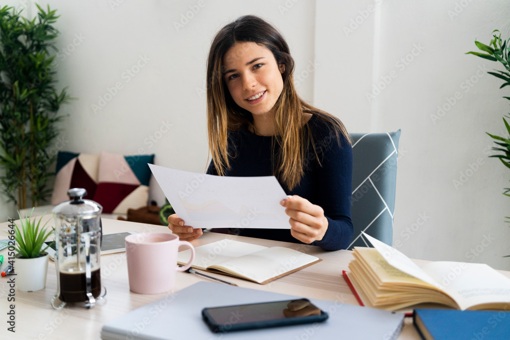 Smiling female freelance worker holding paper while sitting on chair at home Smiling female freelance worker holding paper while sitting on chair at home