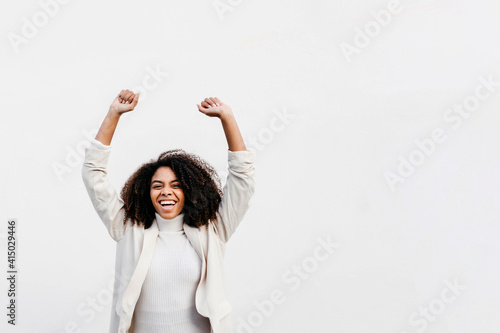 Cheerful afro woman with arms raised screaming while standing against white wall photo