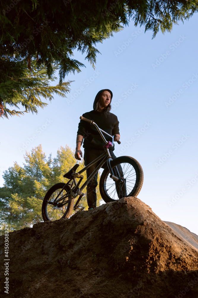 Caucasian white boy with long hair posing with his BMX bike on a sand mountain with trees and blue sky sunny day black sweatshirt