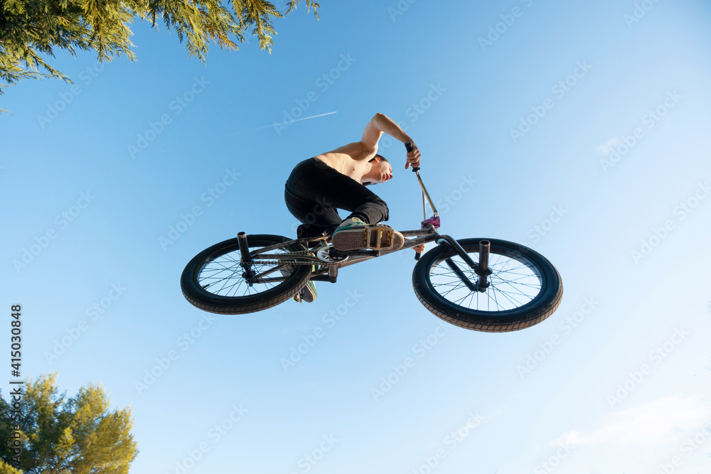 Obraz premium Caucasian white boy jumping with long hair shirtless with BMX bike on a mountain in the field with sand and trees blue sky sunny day