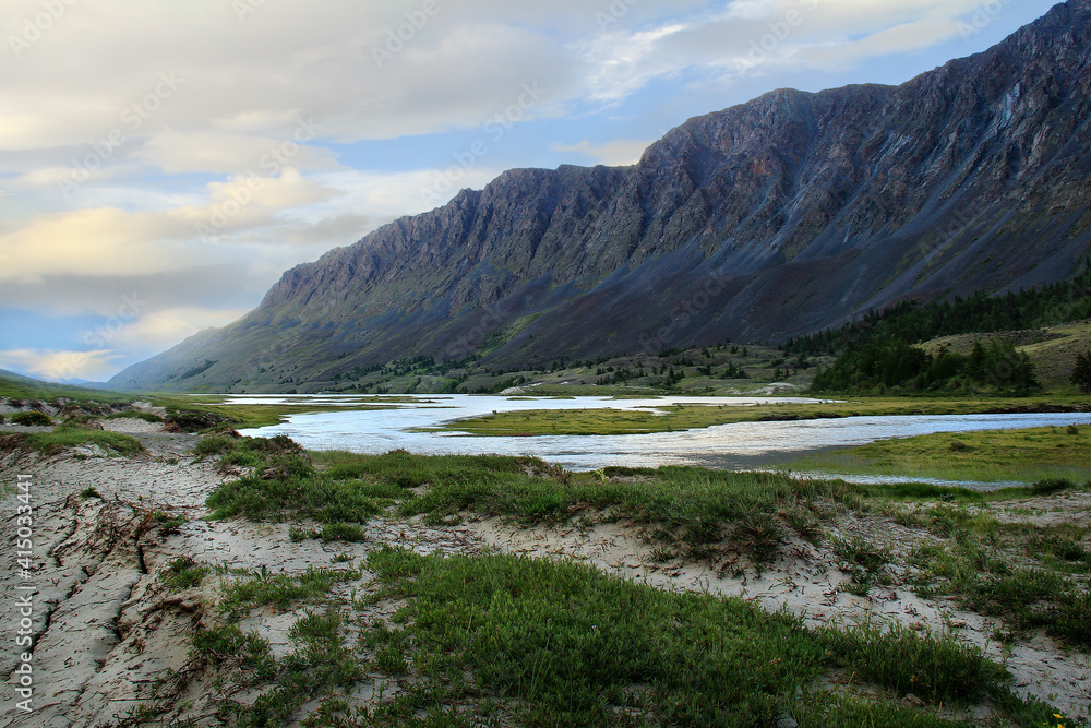 Alpine valley in Altai with the Ak-Kol river, a huge mountain range with rocky peaks, a clay river bank with baiting and forest, a sky with clouds at sunset in summer