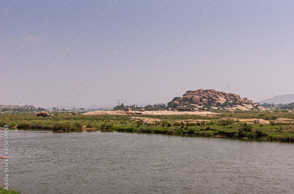 Anegundi, Karnataka, India - November 9, 2013: Gray Tungabhadra river. Wide landscape with Sooryanarayana Temple complex on its green island under light blue sky. Brown rocky hill in back.