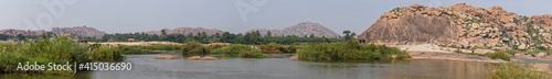 Anegundi, Karnataka, India - November 9, 2013: Wide Panorama shot over Gray Tungabhadra river with green covered islets and brown rocky hills on side and horizon under light blue sky.
