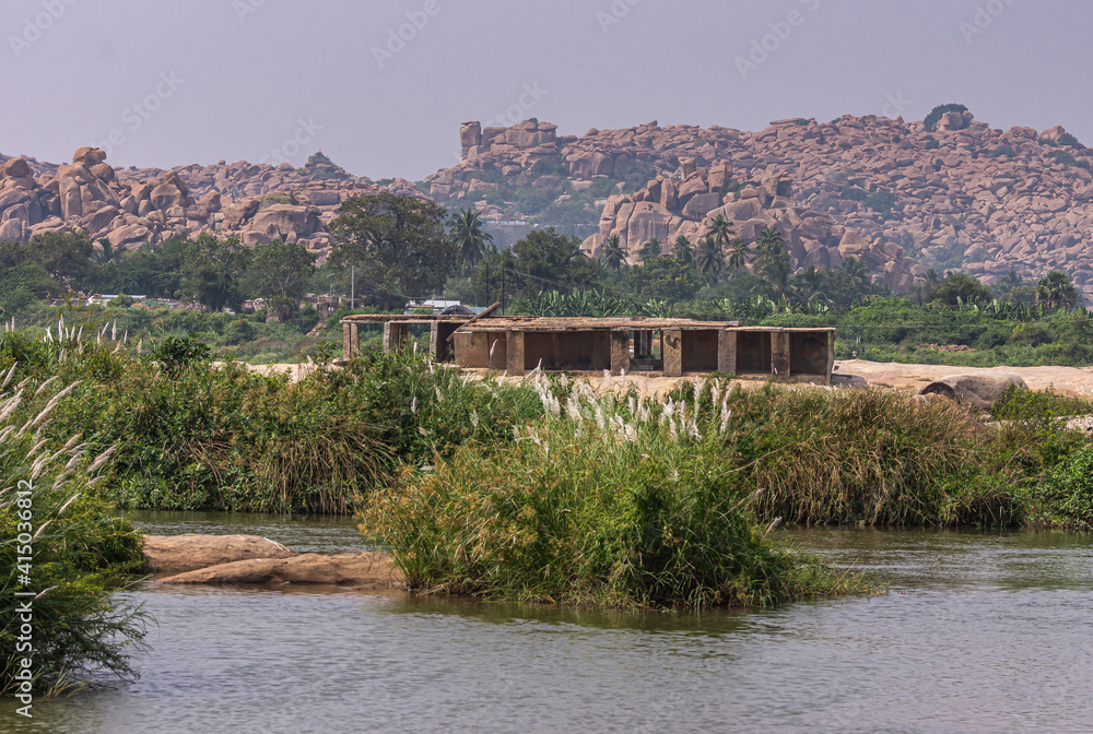 Anegundi, Karnataka, India - November 9, 2013: Gray Tungabhadra river. Building ruins adjacent to Sooryanarayana Temple complex on its green island under light blue sky. Brown rocky hill in back.