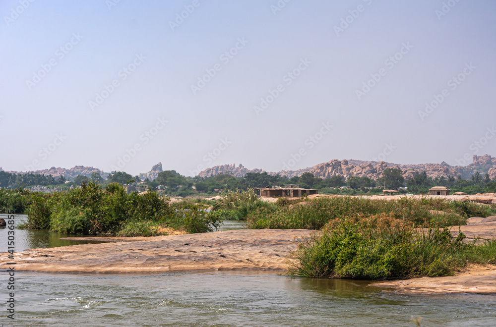 Anegundi, Karnataka, India - November 9, 2013: Sooryanarayana Temple island on gray Tungabhadra river under light blue sky. Green weeds on brown rocks.